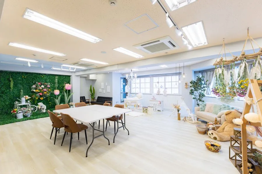 Moff room三宮店の室内全景。中央に白いテーブルと椅子6脚、左に花を飾ったグリーンの壁、右にドライフラワーと大きなくまのぬいぐるみを置いたソファスペースがある明るい完全個室